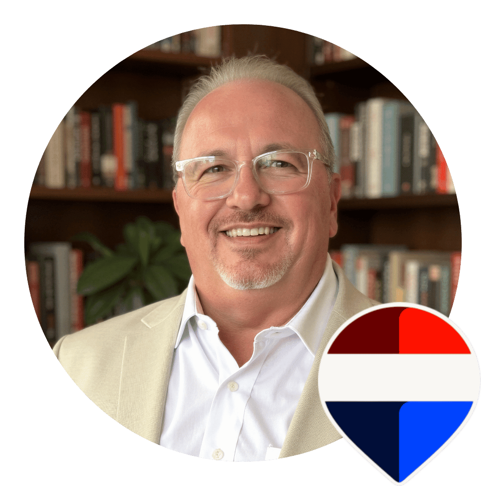 Smiling man in glasses and beige blazer against bookshelf background.